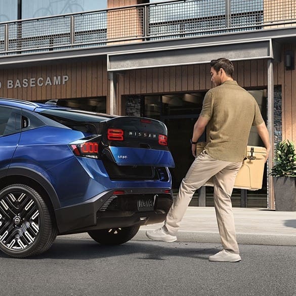 A man stands near a blue Nissan Leaf, preparing to open the trunk while holding a tan cooler bag, with a modern building in the background.