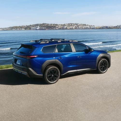 A sleek blue Toyota bZ Woodland is parked by a scenic waterfront, under a clear sky. The vehicle's modern design contrasts with the serene water and distant shore.