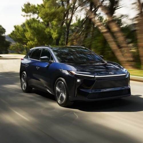 A sleek, dark blue Toyota bZ drives swiftly on a sunny road flanked by blurred trees, conveying motion and modernity in a serene, natural setting.