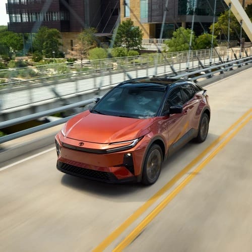 A sleek, red Toyota C-HR drives on a bridge over a river, surrounded by lush greenery and modern buildings under a clear, sunny sky.