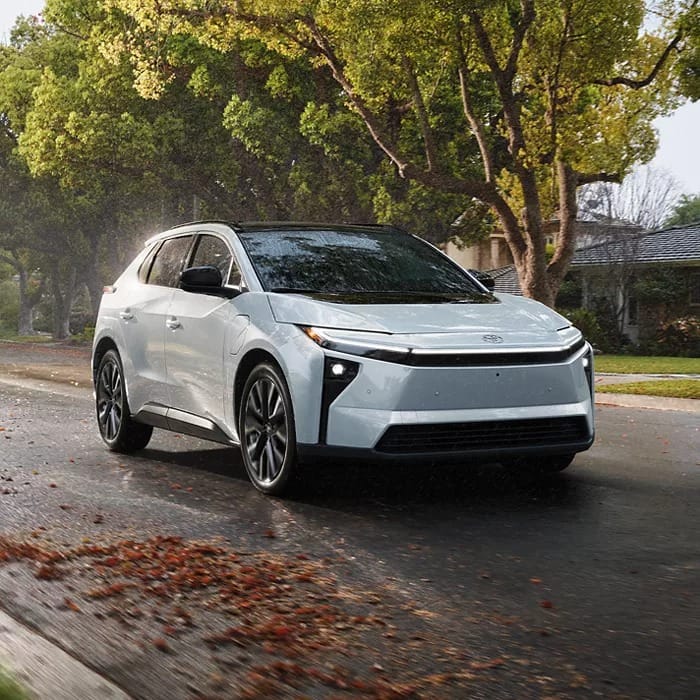 A sleek white Toyota bZ drives on a wet road lined with fallen leaves. Tall trees and a suburban house are in the background, conveying a serene, rainy day.