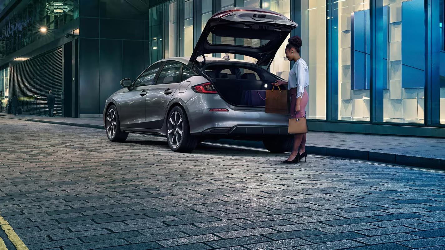 A woman stands beside an open 2026 Honda Civic Hatchback Hybrid trunk on a city street at night, holding a handbag. The scene is illuminated by modern glass buildings, evoking a sleek, urban ambiance.