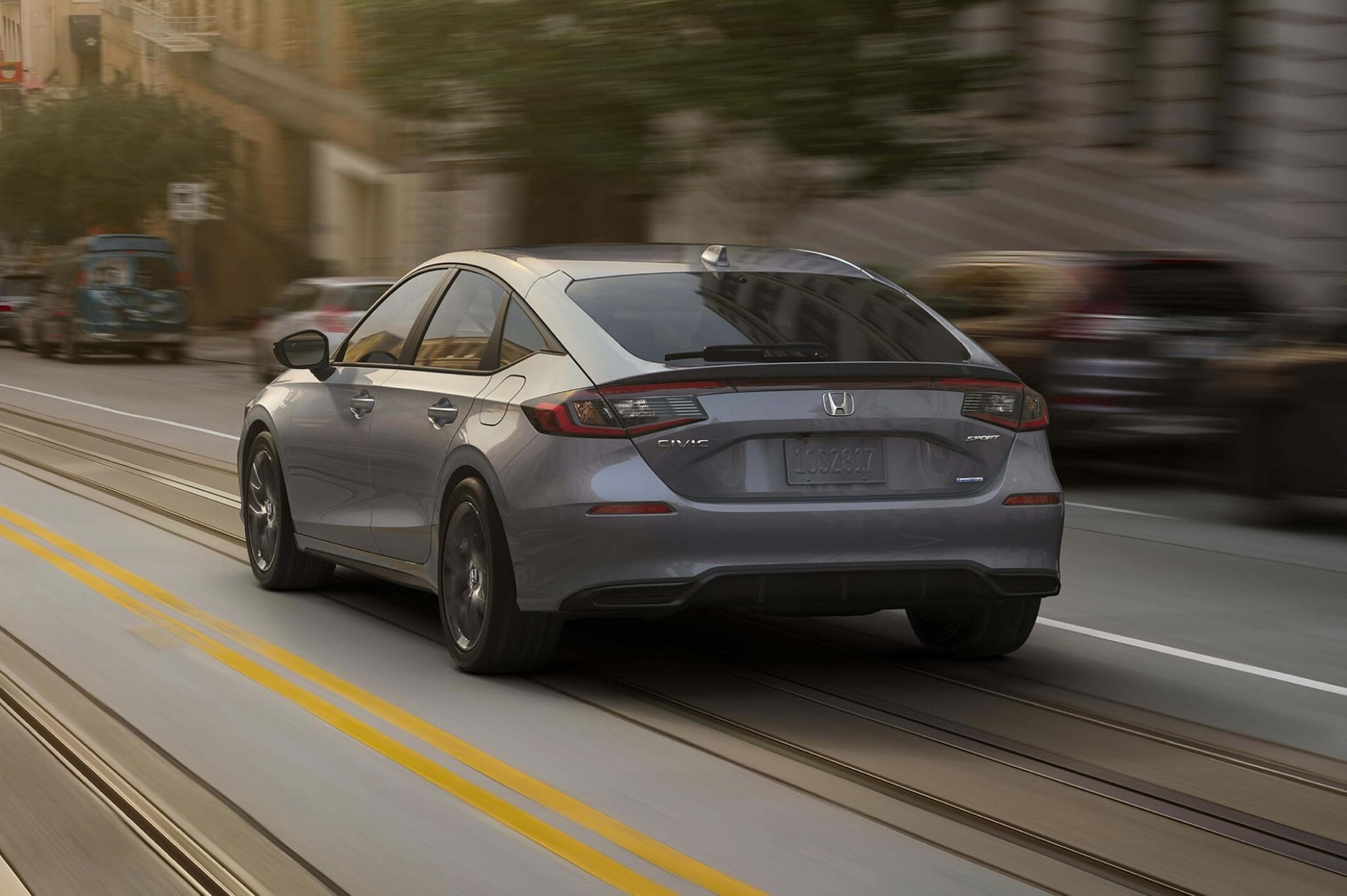 A sleek gray 2026 Honda Civic Hatchback drives swiftly down a city street, with blurred buildings in the background, conveying motion and urban energy.