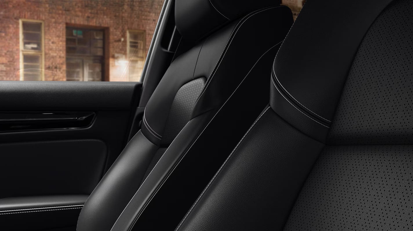 Close-up of a 2026 Honda Civic Hatchback Hybrid interior, featuring sleek black leather seats with white stitching. A brick wall is visible through the window, adding contrast.