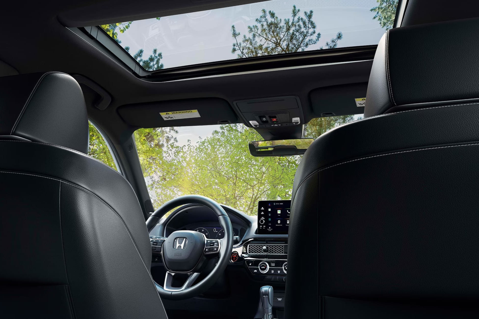 View from the back seat of a 2026 Honda Civic Hatchback interior with black leather, featuring a sunroof and a touchscreen display. Trees visible through the windows.