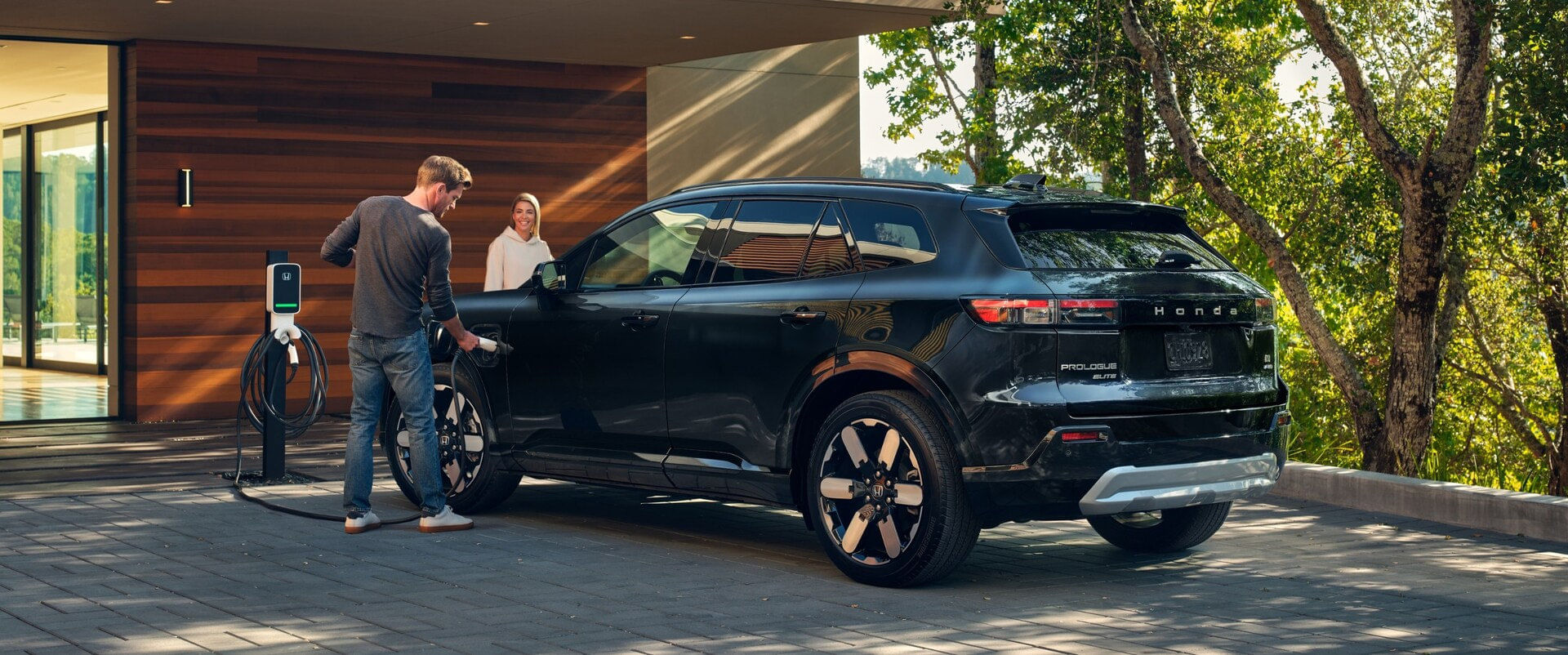 A man plugs a charger into the front fender port of a black 2025 Honda Prologue Elite SUV parked in the driveway of a modern home, while a woman smiles from the passenger seat. A wall-mounted charger is visible
