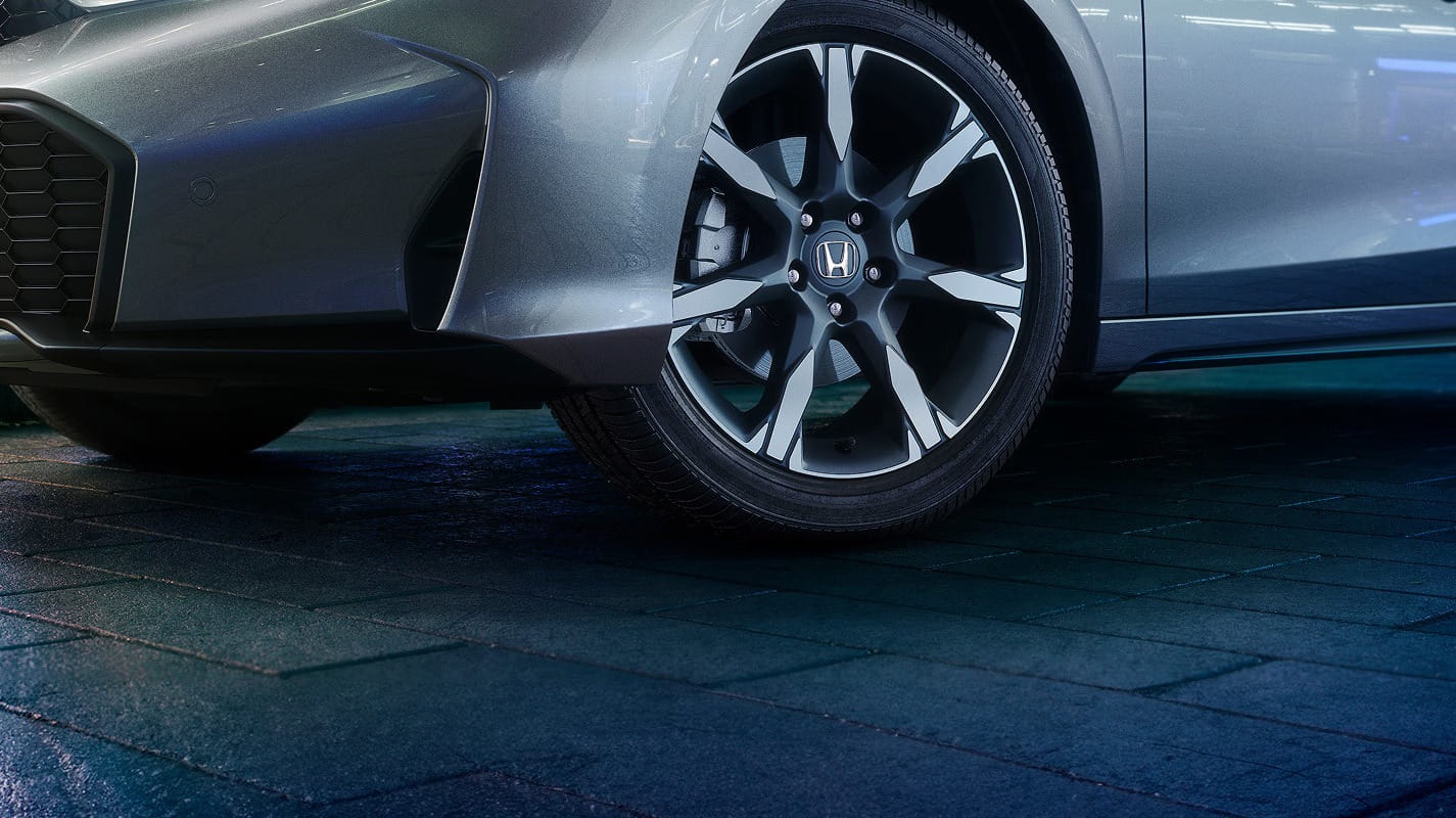 Close-up of a 2026 Honda Civic Hatchback Hybrid front tire and wheel on a tiled, illuminated floor. The setting conveys modernity and sophistication.