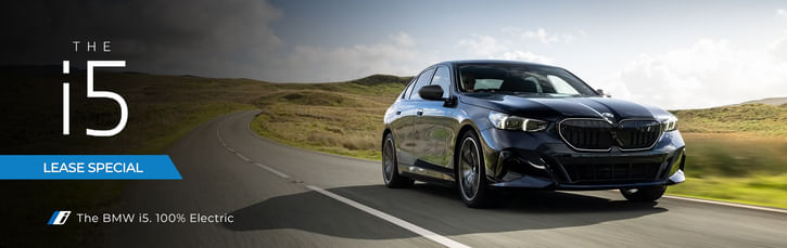 A sleek BMW i5 electric sedan drives along a winding road, surrounded by lush green hills under a clear sky.