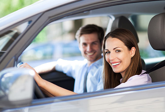 A smiling woman sitting behind the wheel of a gray car, next to her sits a smiling man