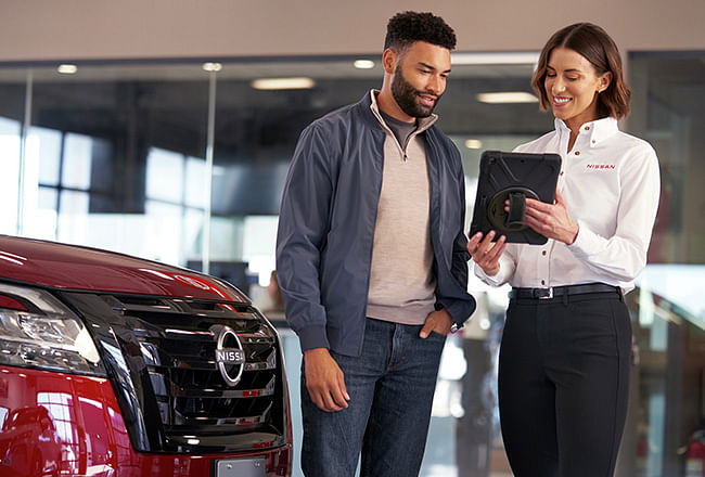 A saleswoman shows a client something on a tablet, a red Nissan is next to it.