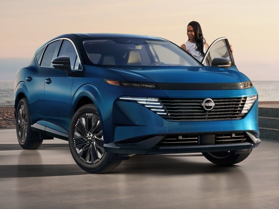 A sleek teal 2026 Nissan Murano is parked by the ocean at sunset, showcasing its modern design and innovative front grille.