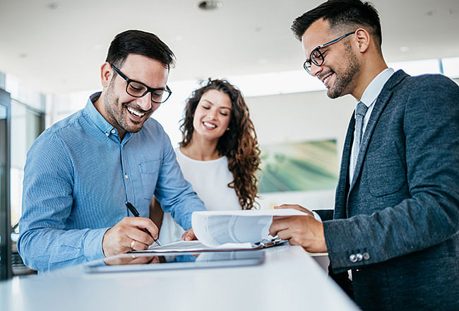 A smiling man signs documents, a salesman stands across from him