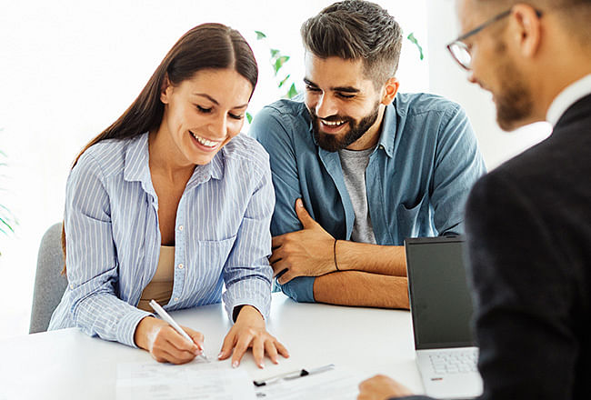 A smiling woman signing documents, a smiling man sits next to her, opposite is a salesman