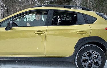 Side of a yellow car with a woman behind the wheel and a brown and white dog in the back