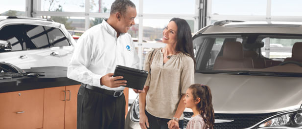 Service advisor in a Mopar-branded shirt smiling and talking with a female customer and her young daughter inside a car dealership service area, with vehicles visible in the background.