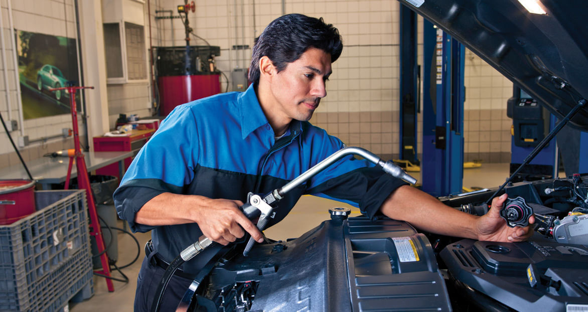 Auto technician in a Mopar uniform performing an oil change, holding a hose and unscrewing the oil cap under the hood of a vehicle in a professional service garage