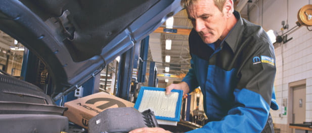 Automotive technician inspecting and replacing an engine air filter under the hood of a vehicle inside a well-lit service garage. The technician is wearing a Mopar-branded uniform.