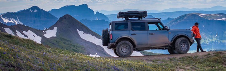 A person in a red jacket stands beside a rugged blue off-road SUV on a mountain trail, with sweeping, snow-patched peaks under a clear sky.