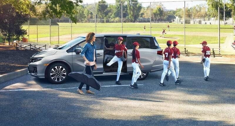 The 2026 Honda Odyssey Elite in Solar Silver Metallic  parked in parking lot with baseball field in the background and several kids exiting the vehicle