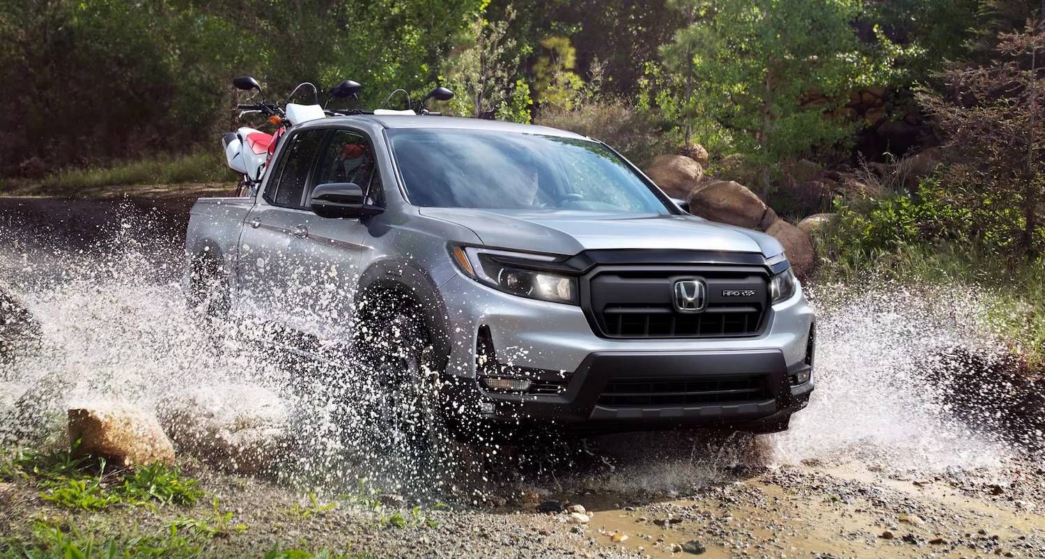 The 2025 Honda Ridgeline Sport in Solar Silver Metallic driving through water with dirt bikes in back