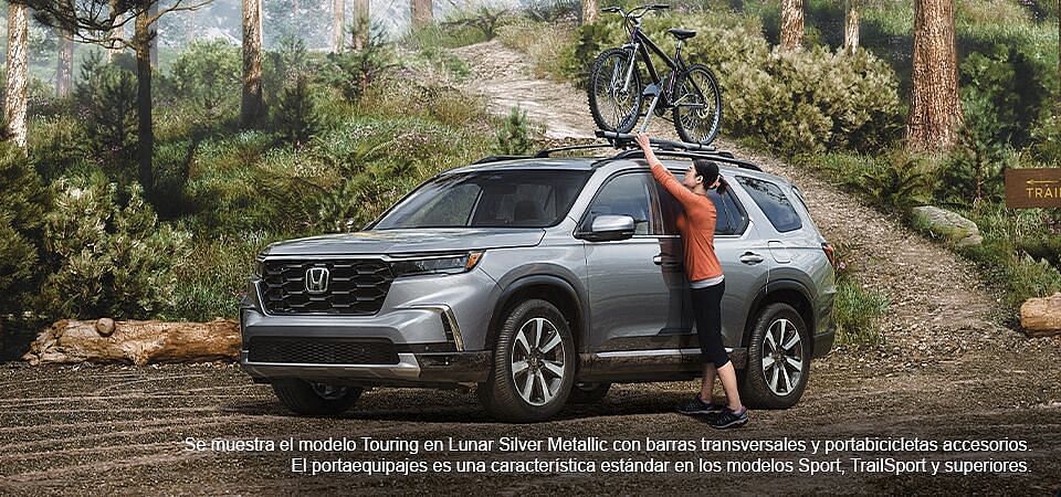 Mujer colocando una bicicleta en el Honda Pilot 2023 en color Lunar Silver metalizado con barras transversales accesorias y enganche para bicicleta en un aparcamiento forestal.