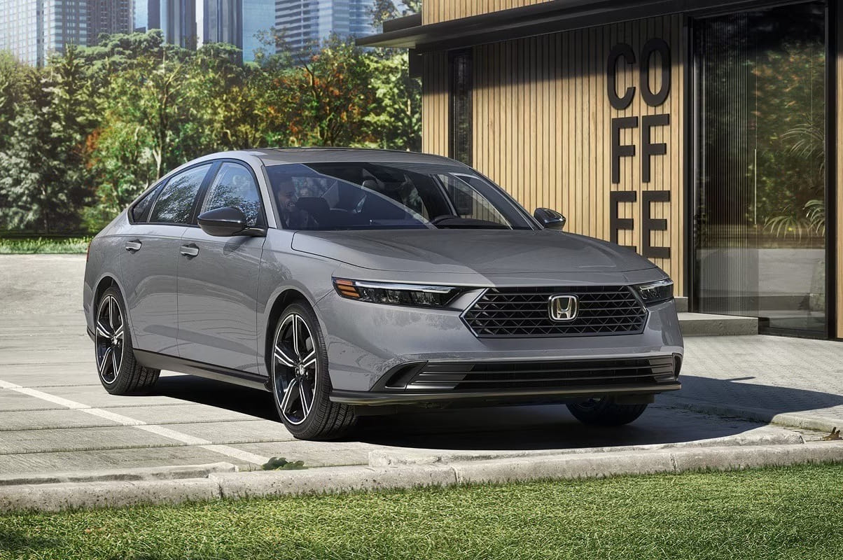 A sleek gray 2026 Honda Accord Hybrid is parked in front of a modern building with “COFFEE” on the window. Trees and skyscrapers are visible in the background.