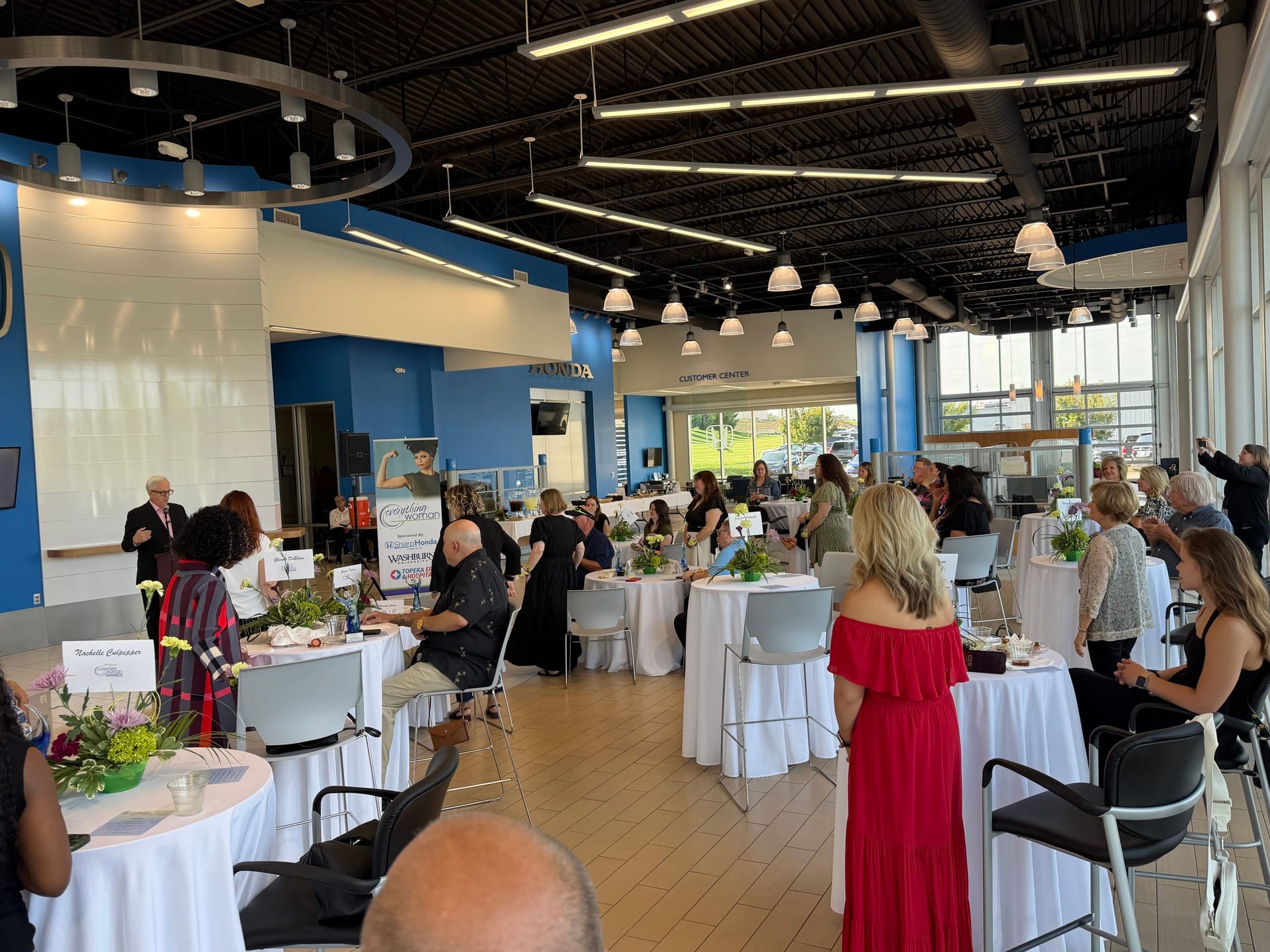 Guests gather at a bright, modern event venue, mingling around white-clothed tables with floral centerpieces in a lively atmosphere.