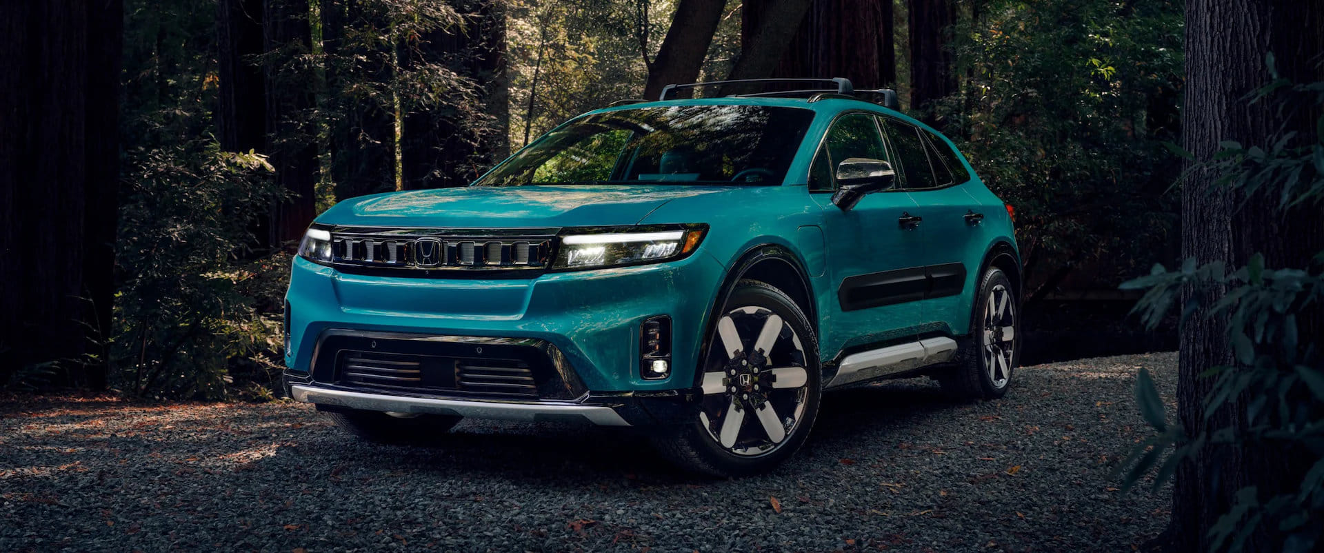 Teal 2025 Honda Prologue SUV with a roof rack, parked on a gravel path in a shaded forest setting with tall trees