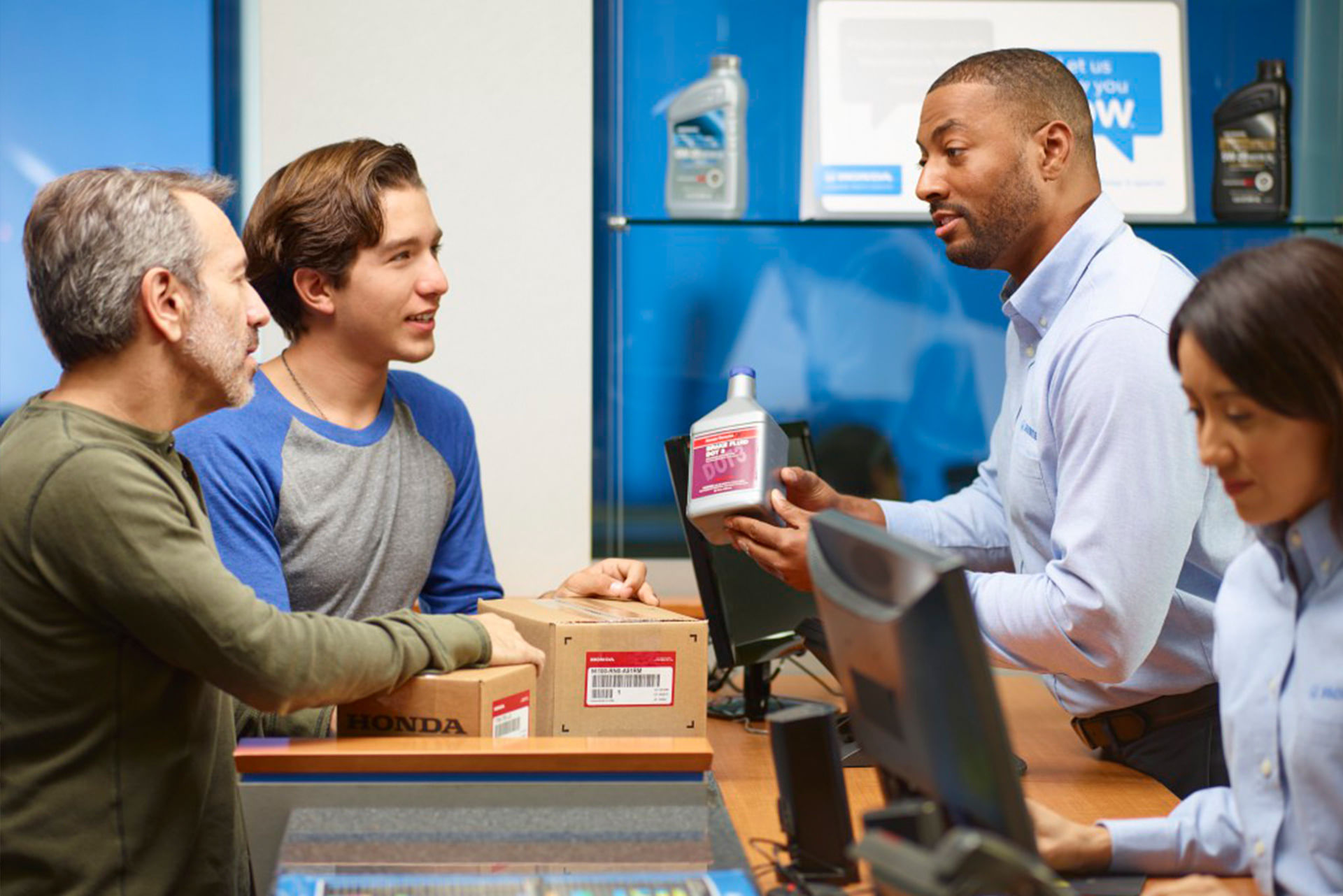 Two customers standing at a parts counter speaking with a Honda employee who is holding a bottle of transmission fluid, while another employee works at a computer in the background.