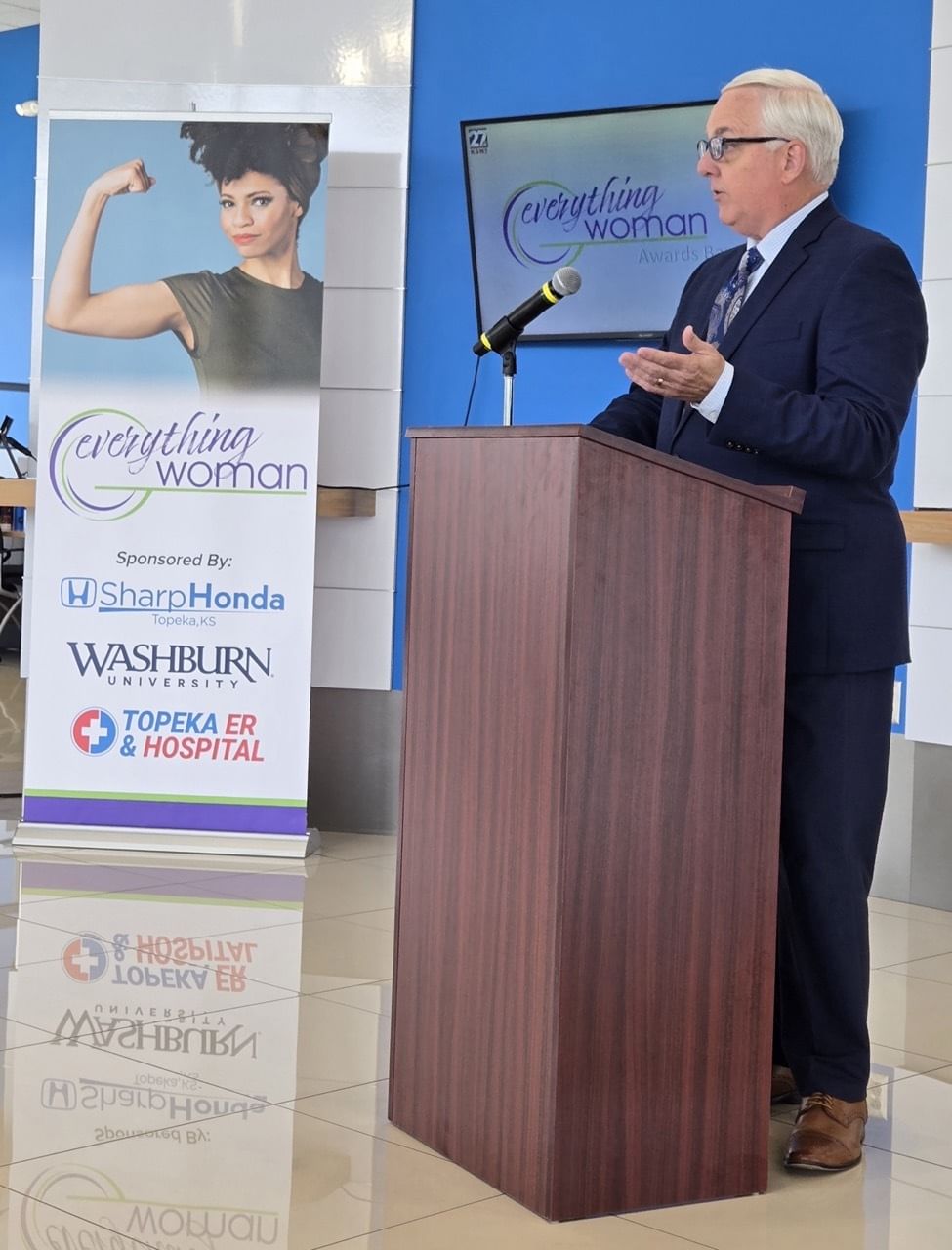 Man in suit addresses attendees from podium at 'Everything Woman' event.