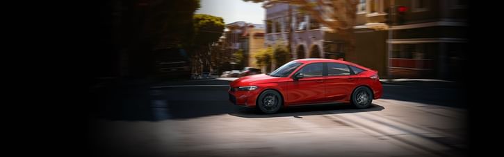 A sporty red 2025 Honda Accord Hybrid speeds around a city corner, showcased against a blurred urban backdrop on a sunny day.