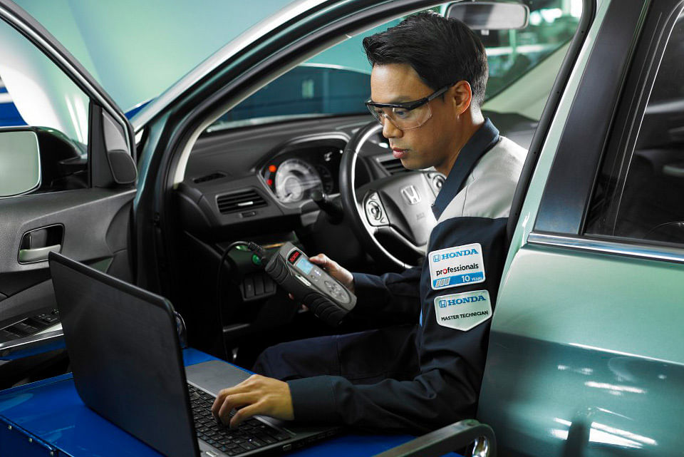 A professional technician in a uniform uses a diagnostic tool and a laptop inside a car with an open door, focusing intently on the task.
