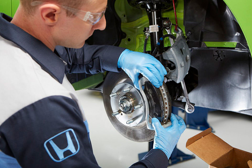 A mechanic wearing gloves and safety goggles installs a brake disc on a vehicle. A cardboard box with tools is nearby, conveying a focused work setting.