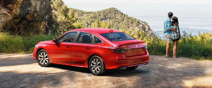 A red Honda Civic sedan is parked on a dirt road at a scenic overlook. To the right, a couple embraces while looking out over a tree-covered hilly landscape towards a large body of water, with a rocky cliff face to the left of the car.