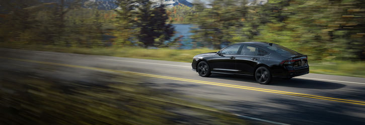 A sleek, black 2025 Honda Accord Hybrid speeds along a scenic road, surrounded by vibrant autumn foliage and a serene lake in the background.