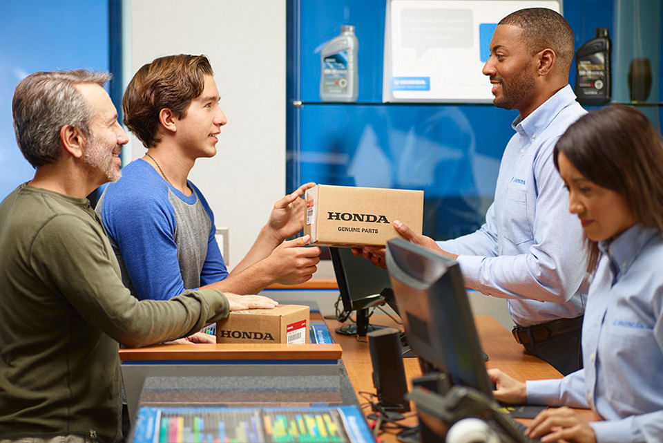 Two customers at a Honda service desk receive genuine parts from smiling staff in blue uniforms, conveying a friendly and professional atmosphere.