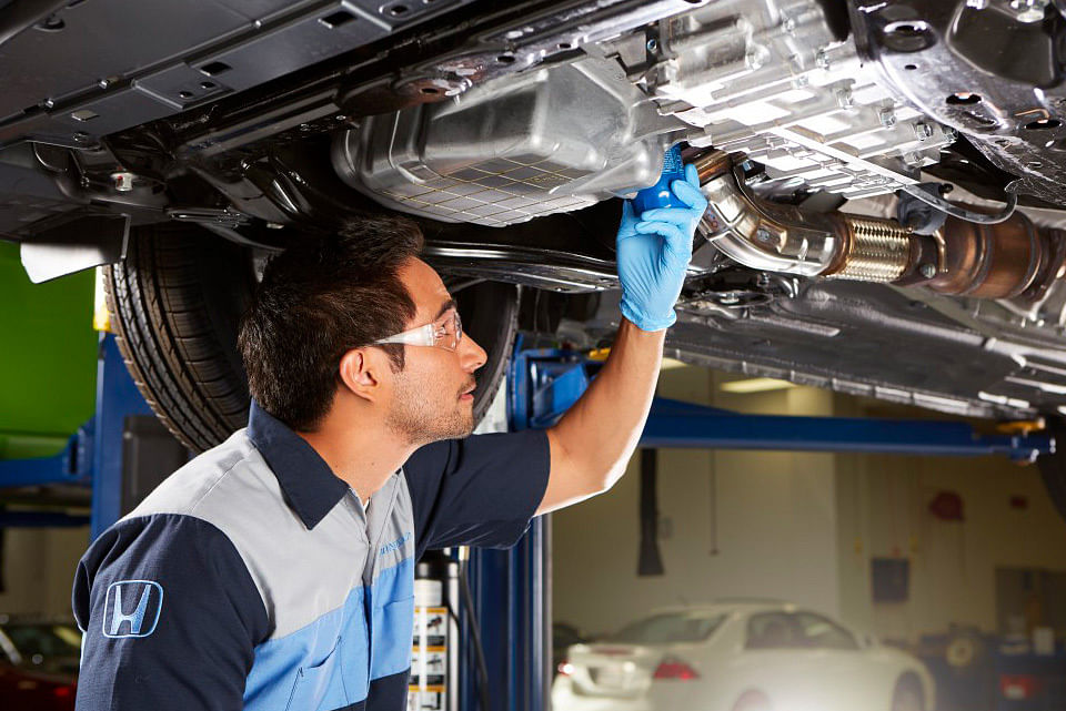 A mechanic in safety glasses and gloves inspects the underside of a car in a well-lit garage, conveying focus and professionalism.