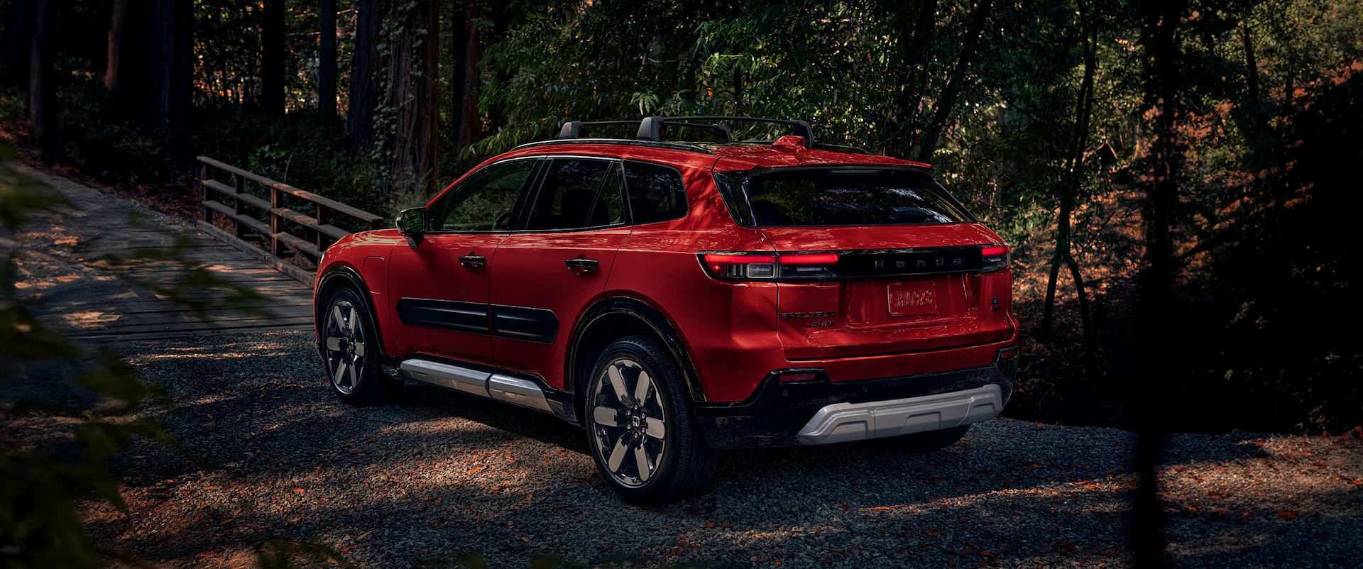 Red 2025 Honda Prologue Elite SUV with a roof rack, parked on a gravel path near a wooden footbridge in a sun-dappled forest