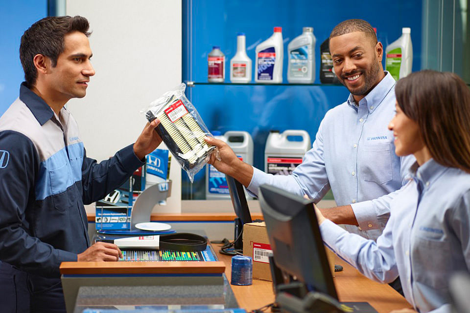 Three people in an auto parts store interact warmly. A worker hands a car filter to a customer over the counter. Shelves of products line the background.