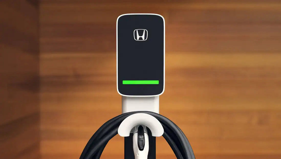 Close-up of a white, wall-mounted Honda EV home charging station against a wood-paneled wall, displaying a green charging status light bar.