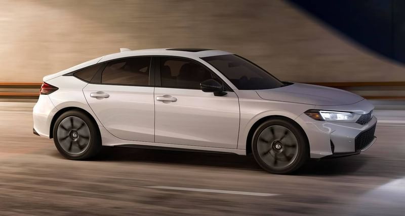 The 2025 Honda Civic Hatchback Sport Touring Hybrid in Platinum White Pearl driving in urban environment