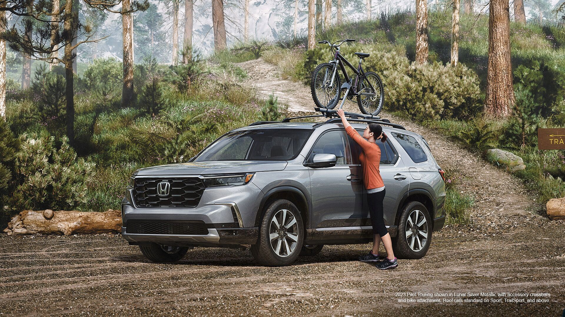 Woman placing bicycle on 2023 Honda Pilot shown in Lunar Silver metallic with accessory crossbars and bike attachment standing on a forest parking.