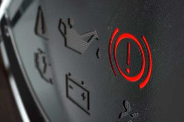 Close-up of a dark vehicle dashboard with a glowing red brake warning symbol—an exclamation mark inside a circle—beside dim icons, conveying an alert.