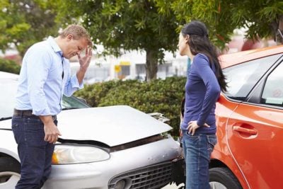 A worried man and woman inspect a dented white car beside an orange vehicle after a minor collision, appearing concerned at the roadside.