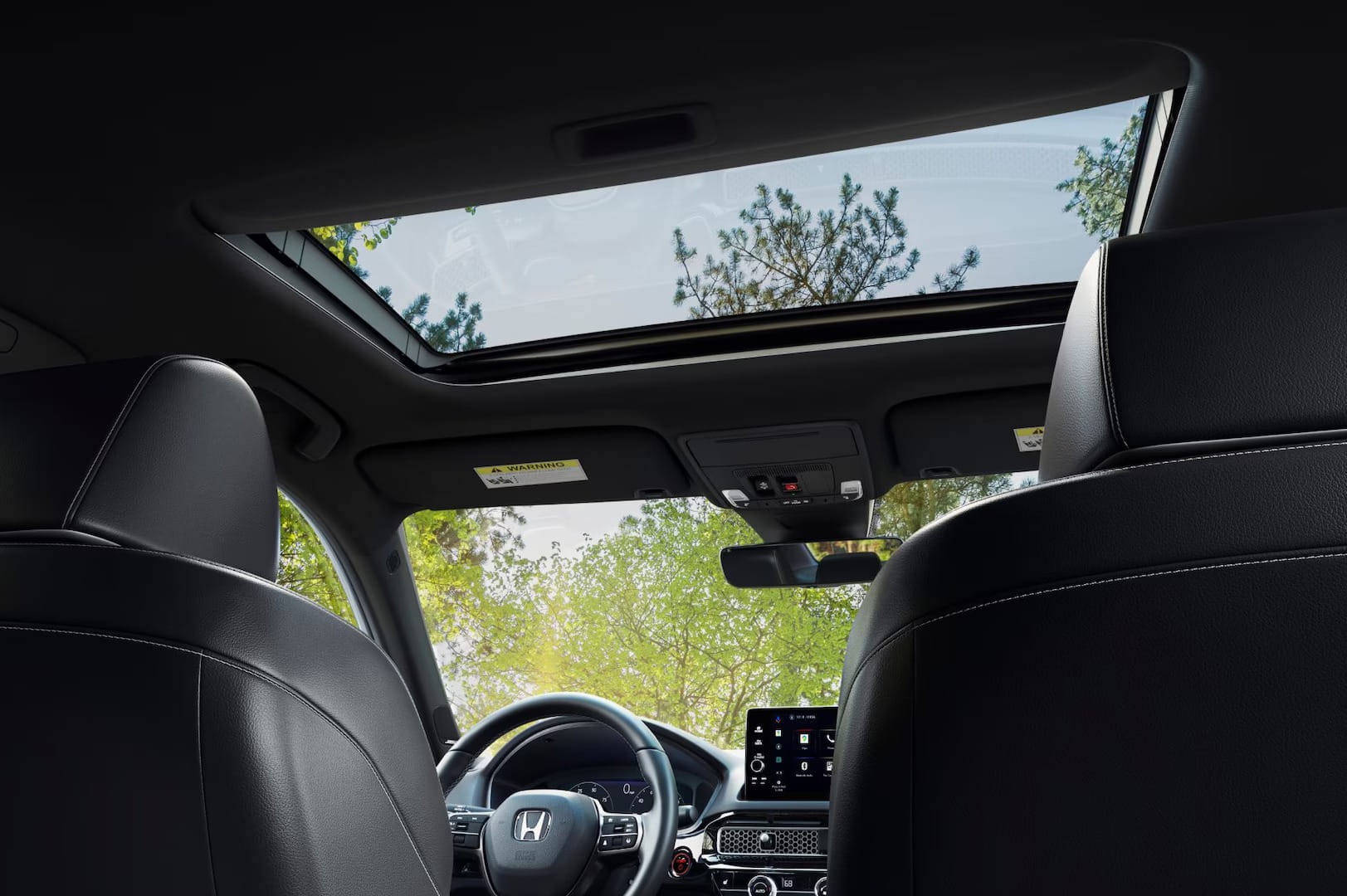 Interior view of a 2026 Honda Civic Sedan Hybrid with black leather seats, a visible steering wheel featuring a Honda emblem, and a sunroof showing green trees and blue sky.