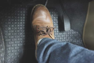 Close-up of a brown leather shoe and blue-jean-clad leg resting on a car’s textured floor mat beside the pedals, evoking a calm moment in the driver’s seat.