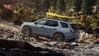 Un Honda Pilot TrailSport 2023 en color Sonic Gray Pearl con barras transversales accesorias y enganche para kayak circulando por un arroyo forestal.