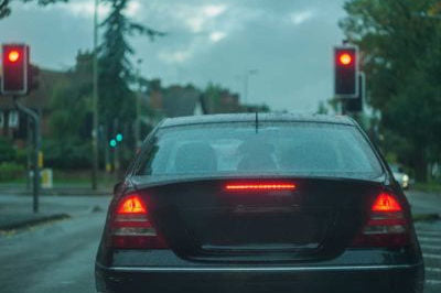 A dark sedan waits at a rain-slick intersection beneath red traffic lights, its brake lights glowing against a misty, overcast city street.