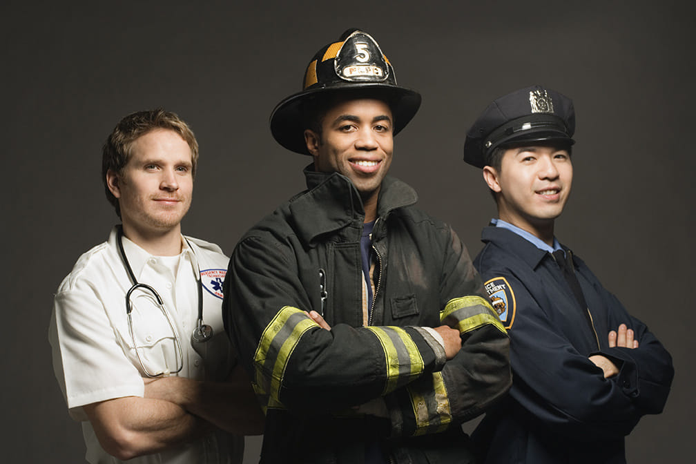 Studio portrait of three first responders standing together: a male paramedic with a stethoscope, a male firefighter in turnout gear and helmet, and a male police officer in uniform.