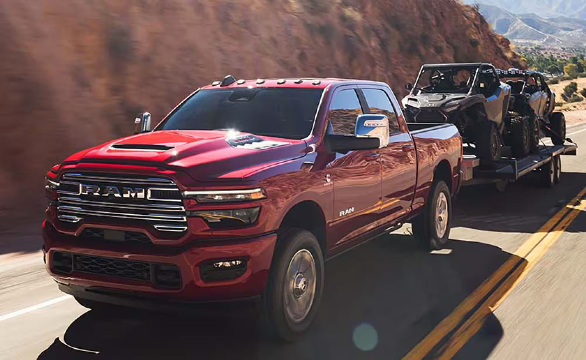 A red 2025 Ram 2500 Laramie 4x4 Crew Cab traveling on a highway in the mountains, while towing two ATVs.